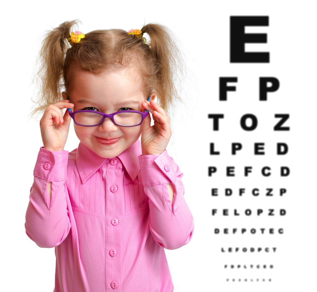 Smiling,Girl,Putting,On,Glasses,With,Blurry,Eye,Chart,Behind